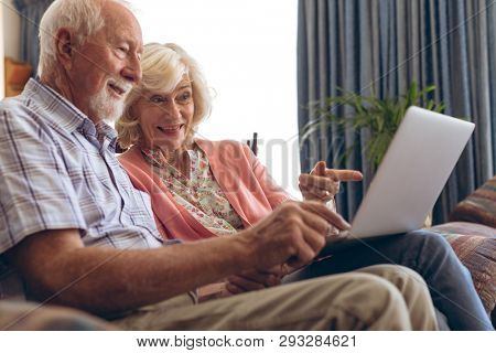 Side view of senior Caucasian couple interacting with each other while using laptop at retirement home. Couple sits on sofa while pointing with their fingers towards the screen of the laptop.