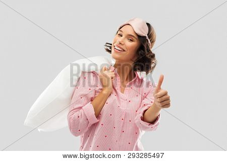 people and bedtime concept - happy young woman in pajama and eye sleeping mask with pillow over shoulder showing thumbs up over grey background