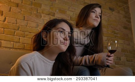 Closeup Side View Portrait Of Two Young Pretty Women Watching Tv And Drinking Wine In A Cozy Apartme