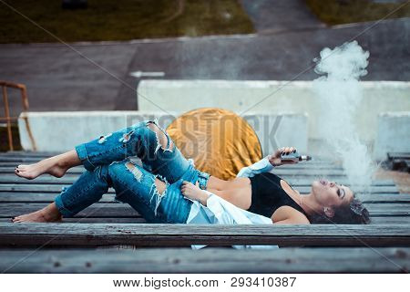 Young Sexy Woman Lying On The Floor And Smoking Vaping E-cigarette. She's Wearing A Top And A White 