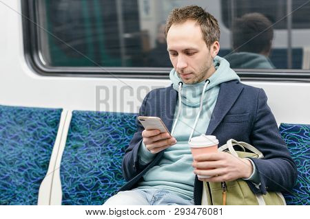 Handsome Man In Blue Coat And Turquoise Hoodie Is Sitting In A Subway Train, Drinking Coffee And Sur