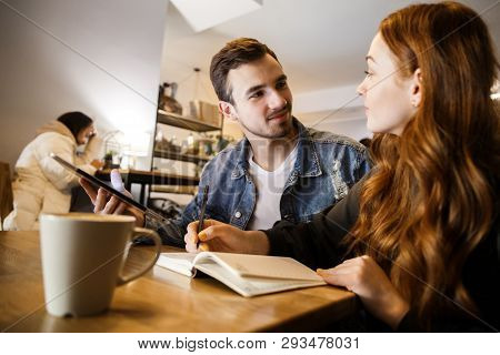 A Young Pair Is Sitting At The Cafe. They Are Looking At Each Other. A Young Man Is Holding A Tablet