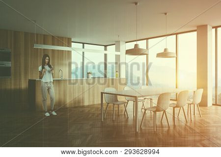 Woman With Smartphone Standing In Modern Wooden Kitchen With Wooden Countertops, White Dining Table 