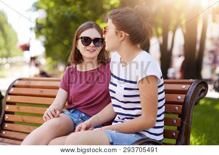 Two Creative Girls Talk And Laugh While Sitting On Bench Outdoors. Young And Funloving Friends Share