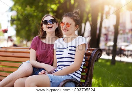A Portrait Of Two Attractive Young Friends Who Have Rest On Wooden Bench In Local Park After Long Wa