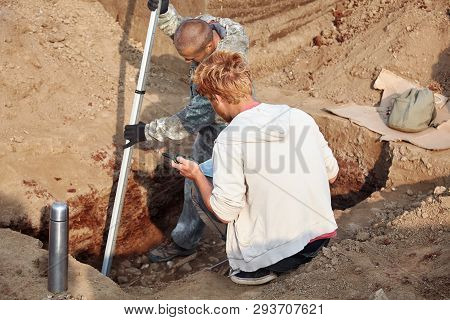 Two Men At Work Outdoors, In Digger Process. Archaeological Excavations. One Archaeologist With A Mo