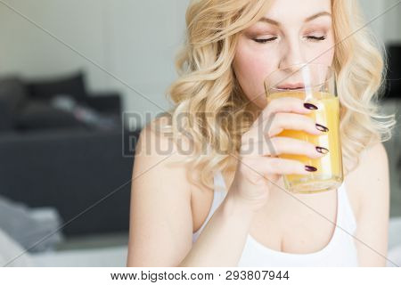 Attractive Blonde Woman Sits On The Edge Of The Bed At Home And Holds A Stocker With Orange Juice In