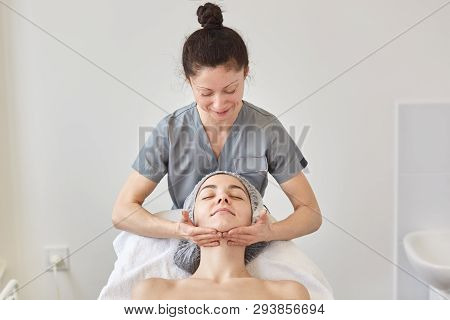 Cosmetologist Wears Gray Gown Massaging Patient Face. Young Woman With Naked Shoulders And Dresses M