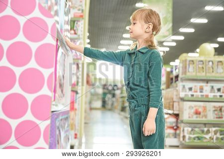 Pretty, Cute Girl Standing Near Shelves With Toys, Looking And Choosing Goods. Big Choice Of Toys Fo