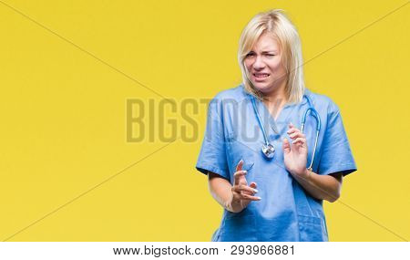 Young beautiful blonde doctor woman wearing medical uniform over isolated background disgusted expression, displeased and fearful doing disgust face because aversion reaction. With hands raised