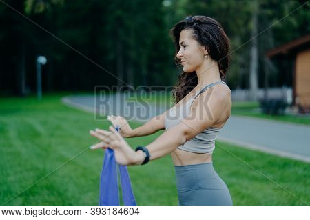 Profile Shot Of Sporty Slim Woman Holds Resistance Band, Has Fitness Training, Dressed In Sportswear