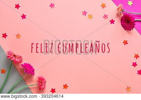 Feliz Cumpleanos Means Happy Birthday In Spanish Language. Flat Lay, Gerbera Flowers On Pink Paper