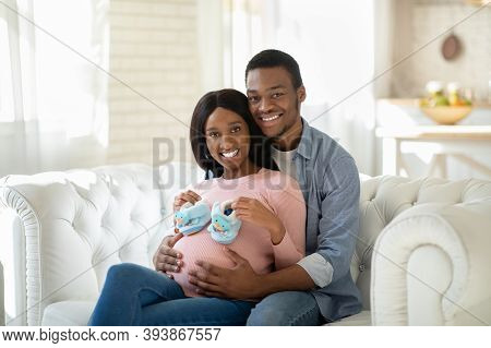 Black Pregnant Woman With Tiny Baby Boties And Her Happy Husband Hugging On Sofa At Home. Future Mom
