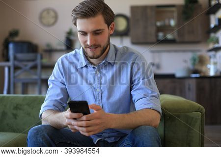 Attractive Young Man Relaxing On A Couch At Home And Using Mobile Phone For Cheking Social Nets.