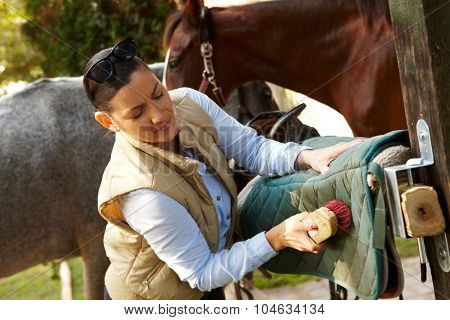 Young woman cleaning saddlery outdoors, brushing saddle-cloth.
