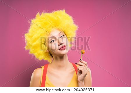 A funny girl in a yellow wig looks thoughtfully to the right and eats chupa-chups. Tired Clown