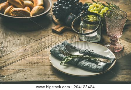 Fall holiday table decoration setting. Plate with linen napkin, fork and spoon, candle, fresh fruits on serving board, bread buns, olive tree branch over wooden background, selective focus, copy space