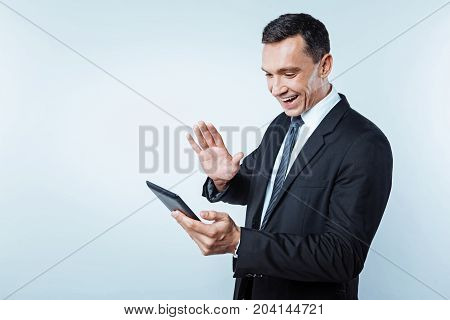 Hello there. Waist up shot of a beaming mature businessman waiving to somebody while focusing his attention on a screen of a tablet computer during a video call conversation.