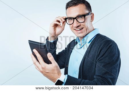 No time for worries. Educated man wearing a smart casual attire touching his glasses while looking into the camera with a broad smile on his face with a digital tablet in hand.