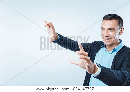 Latest technologies. Cheerful guy in smart casual grinning broadly while lifting his hands and pretending to touch a transparent screen.