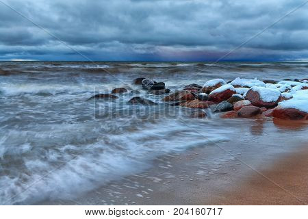 Winter storm with waves on the Baltic in the late evening against a background of stones with snow on a sandy beach