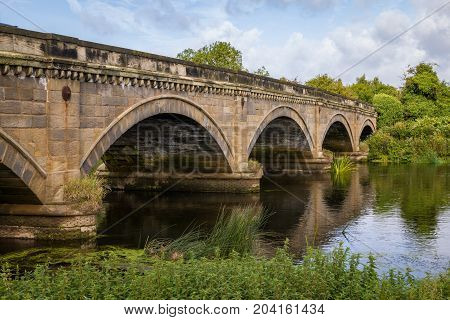 Stone Bridge over The River Trent between Repton and Willington Derbyshire. Showing the river cloudy sunny blue sky green folage foreground reflection of bridge in water.