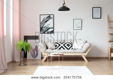 Pillows on beige sofa against wall with posters in simple living room with copper table and fern