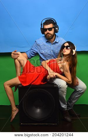 Music and leisure concept. Man with beard wears headphones sunglasses. Couple in love lies on loudspeaker on blue and green background. Guy with beard and lady with serious faces enjoy music