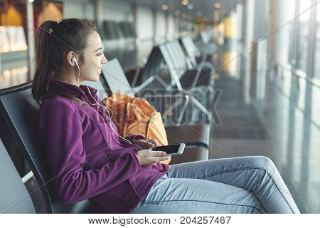 Girl at the airport window sits on the banch and listening to music on headphones from the phone