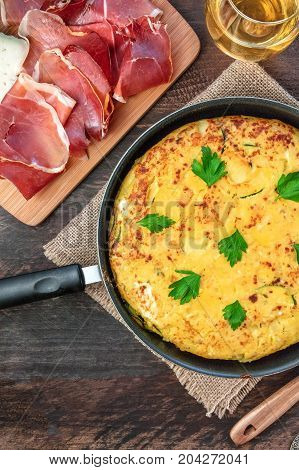 An overhead photo of a Spanish tortilla with potatoes and zucchinis, shot from above on a dark rustic texture in a tortillera, with slices of jamon, and a glass of white wine, with a place for text