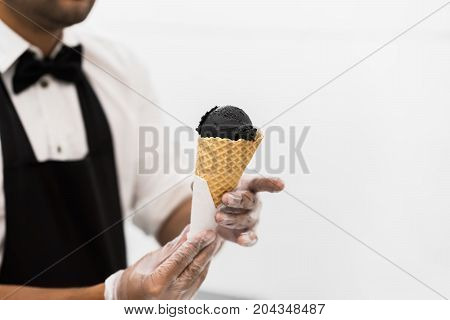 Handsome young waiter in elegant classic suit serving trendy dessert, black ice cream in traditional portioned waffle cone. Uninsulated white background, copy space. Real scene. Professional service, catering concept