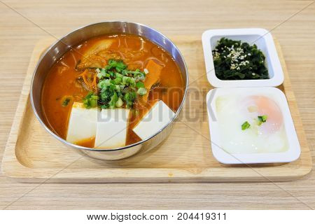 The homemade food set consists of spicy kimchi soup (bacon slice pork white tofu spring onion & cabbage) soft boiled egg (onsen) green seaweed with white sesame serve on wooden tray.