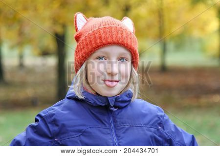 Cheerful girl shows tongue, the child is dressed in a funny knitted warm hat with ears, looks like a fox. Autumn, outdoors portrait.