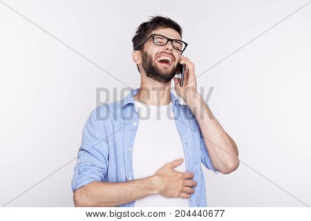 Half profile of attractive young bearded man wearing casual white shirt stylish shades with happy expression and mouth wide open laughing while having a phone conversation using generic smart phone.