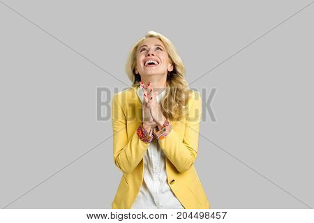 Happy grateful young blond woman. Joyful young lady showing her gratitude clasping her hands and lifts eyes to thew sky over grey background.