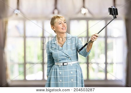Smiling woman taking selfie with monopod. Attractive mature woman taking selfie with monopod on blurred background. Happy white-skin woman with monopod.