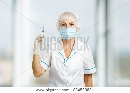 Portrait of nurse with syringe, front view. Close up of female mature doctor holding syringe with injection on blurred background.