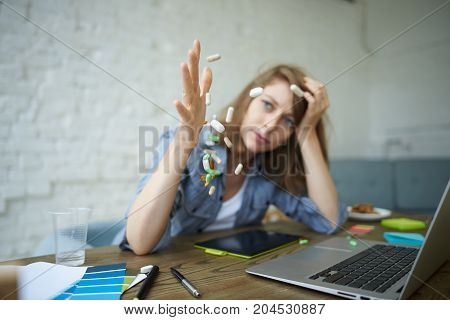 Freeze shot of blue-eyed young European woman freelancer with headache working at home office using notebook pc pouring out assortment of colorful pills on table. Selective focus on hard capsules