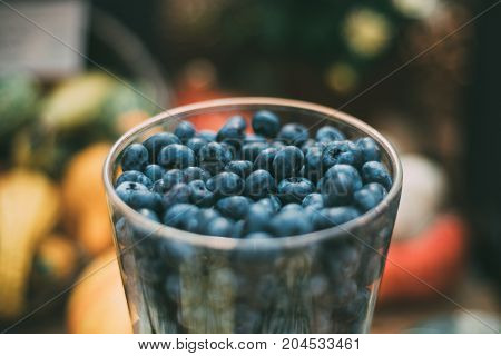Close-up view of glass with fresh raw bog whortleberry or bilberry just recently harvested for Thanksgiven day with colorful spots of defocused vegetables in background; shallow depth of field