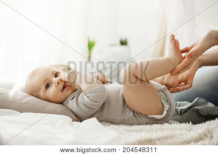 Close up of cute newborn baby lying on bed, looking aside while mother playing and touching his little legs. Baby gnawing finger, his face express happiness and joy