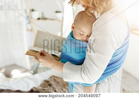 Close up of young mom reading fairy tales for her newborn little son in comfortable light bedroom. Baby falls asleep while she was reading