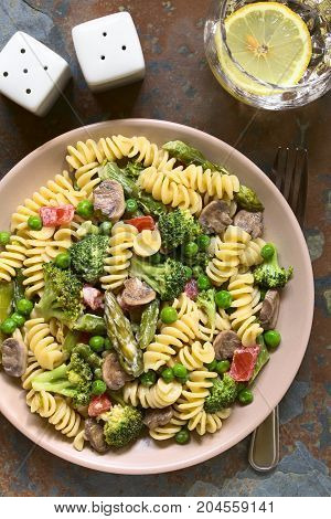 Pasta primavera with green asparagus pea broccoli mushroom and tomato in cream sauce served on plate photographed overhead on slate with natural light