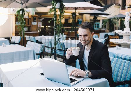 Successful enterpreneur is sitting at the table in a big opened-place restaurant during the short break in a workday. He is drinking coffe and talking with somebody on Skype. The man is happy and smiling