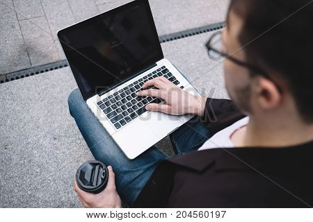 The cut view of a businessman sitting on the concrete ground with the laptop on his knees. He is holding a cup of coffee in his right hand and putting his fingers of his right hand on the keyboard. Close up