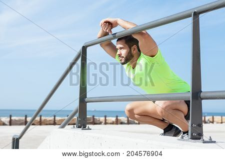 Portrait of young handsome strong man wearing sportswear, looking away, relaxing, sitting on hunkers and leaning on railing at seaside