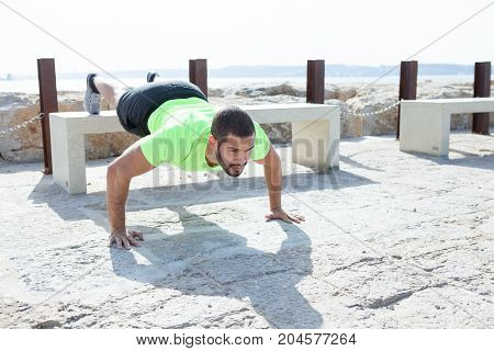 Portrait of focused young strong handsome man wearing sportswear and doing feet elevated push-ups on concrete bench at seaside