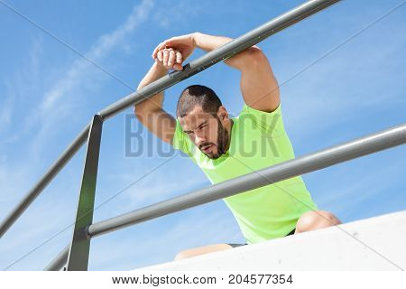 Portrait of young handsome strong man wearing sportswear, looking away, relaxing, sitting on hunkers and leaning on railing at seaside. Low angle view.