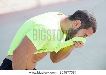 Closeup of young handsome strong man wearing sportswear, relaxing and wiping face with his t-shirt. Side view.