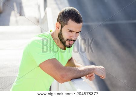 Contemplative handsome fitness trainer being on stroll. Aspirated young man in green t-shirt leaning on railing and looking at road outdoors. Deep in thoughts concept
