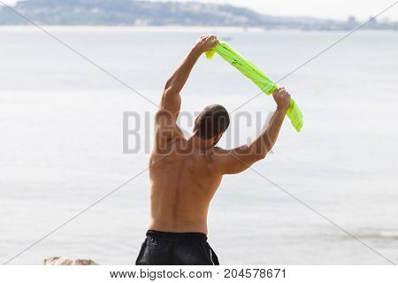 Closeup of shirtless young handsome strong man holding t-shirt, stretching body and doing side bend exercise with sea in background. Back view.